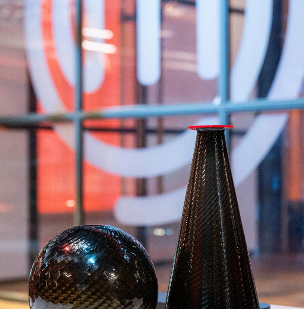 Una sfera in vetro di Murano grigia ed una bottiglia in vetro di murano grigia con bordo rosso a contrasto, appoggiati su un tavolino bianco. Sullo sfondo una vetrata con il logo del programma Masterchef in bianco.