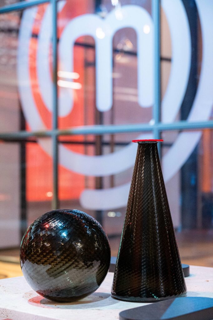 Una sfera in vetro di Murano grigia ed una bottiglia in vetro di murano grigia con bordo rosso a contrasto, appoggiati su un tavolino bianco. Sullo sfondo una vetrata con il logo del programma Masterchef in bianco.